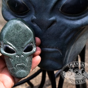 May include: A green stone carving of an alien head with black eyes. The carving is detailed and has a textured surface. The carving is held in a hand.