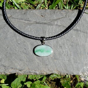 May include: A black braided leather necklace with a silver pendant featuring an oval, green stone. The necklace rests on a gray stone surface, surrounded by green grass and foliage. The pendant is approximately 1 inch in length.