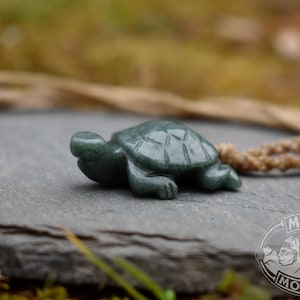 May include: A dark green jade turtle figurine with detailed shell markings. The turtle is lying on a gray stone surface, with a blurred natural background. A natural cord is attached to the turtle.