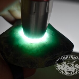 May include: A close-up of a green jade stone being illuminated by a light source. The stone is rough and has a dark green color. The text "MAYAN MOUNTAIN" is visible in the lower right corner of the image.