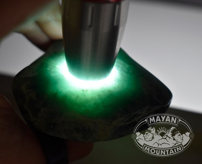 May include: A close-up of a green jade stone being illuminated by a light source. The stone is rough and has a dark green color. The text "MAYAN MOUNTAIN" is visible in the lower right corner of the image.
