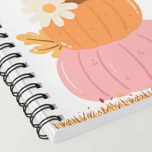 May include: Spiral-bound notebook featuring a fall-themed illustration. The design includes a light orange pumpkin, a pink pumpkin, a white daisy, and autumn leaves. The notebook's edge has a decorative grass border.