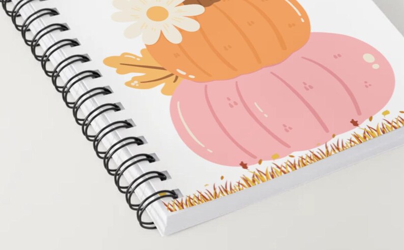 May include: Spiral-bound notebook featuring a fall-themed illustration. The design includes a light orange pumpkin, a pink pumpkin, a white daisy, and autumn leaves. The notebook's edge has a decorative grass border.