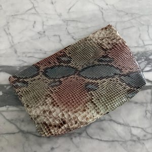 May include: A rectangular clutch bag featuring a textured snakeskin pattern in shades of beige, brown, and muted blue. The surface displays a repeating scale design with a natural gradient, creating a distinct visual pattern across the entire exterior.