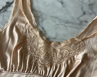May include: A peach-colored satin slip with delicate lace detailing at the neckline. The slip features thin straps and a gathered bodice, creating a soft, flowing silhouette. The lace is a matching peach color, adding a touch of elegance.