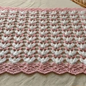May include: A square crochet blanket featuring a repeating pattern of white textured butterfly motifs on a light pink background. The blanket is finished with a wide, scalloped pink crochet border and includes a small circular wooden tag in one corner.