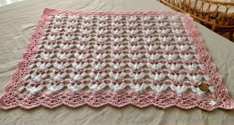 May include: A square crochet blanket featuring a repeating pattern of white textured butterfly motifs on a light pink background. The blanket is finished with a wide, scalloped pink crochet border and includes a small circular wooden tag in one corner.