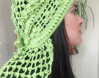 Pode incluir: Um chapéu de crochê verde claro com uma echarpe a condizer. O chapéu tem uma coroa arredondada e uma borda decorativa. A echarpe é presa ao chapéu e cai para o lado. O crochê tem um design aberto e texturizado.