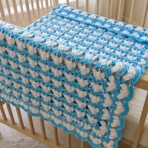 May include: A rectangular crochet blanket featuring a repeating pattern of white and light blue yarn clusters. The blanket has a scalloped light blue border along the edges and is draped over the wooden slats of a crib, displaying a textured finish.
