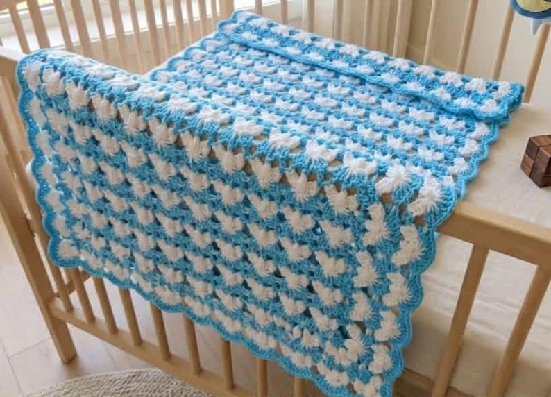 May include: A rectangular crochet blanket featuring a repeating pattern of white and light blue yarn clusters. The blanket has a scalloped light blue border along the edges and is draped over the wooden slats of a crib, displaying a textured finish.