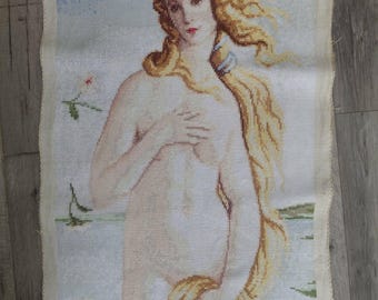 Pode incluir: Uma tapeçaria de ponto cruz que retrata "O Nascimento de Vénus" de Botticelli. A obra de arte apresenta uma figura feminina nua com longos cabelos loiros esvoaçantes, cobrindo o peito com a mão. O fundo inclui um céu azul claro, água e vegetação.