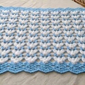 May include: A rectangular crochet blanket featuring a repeating pattern of white, fuzzy butterfly-like shapes on a light blue background. The blanket is finished with a wide, scalloped light blue border and includes a small, round wooden tag.