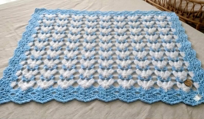 May include: A rectangular crochet blanket featuring a repeating pattern of white, fuzzy butterfly-like shapes on a light blue background. The blanket is finished with a wide, scalloped light blue border and includes a small, round wooden tag.
