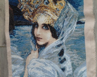 Pode incluir: Uma tapeçaria detalhada retrata uma mulher com olhos azuis e cabelo escuro, usando um adorno de cabeça dourado e azul com joias e um xale azul esvoaçante. A obra de arte tem uma paleta de cores azul e bege, com uma aparência detalhada e texturizada.