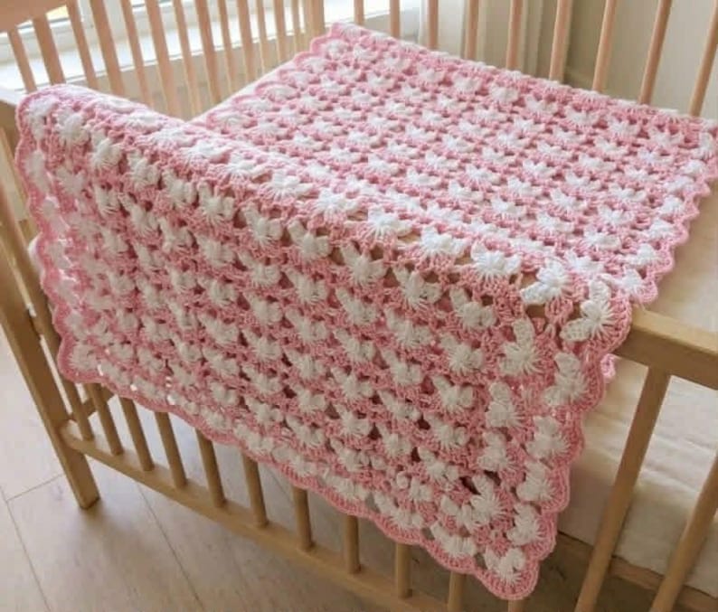 May include: A rectangular crochet blanket featuring a repeating pattern of white and light pink yarn. The textile has a scalloped edge border and an openwork design, showcasing a delicate, interconnected stitch structure throughout the piece.