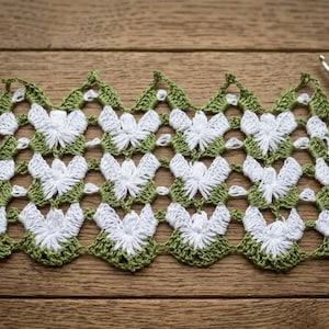 May include: A rectangular piece of crochet lace featuring a repeating pattern of white floral motifs connected by olive green yarn. The lace has a scalloped top and bottom edge. A wooden crochet hook and metal scissors are positioned to the right.