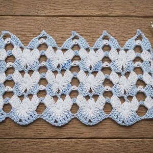 May include: A rectangular piece of crochet lace featuring a repeating chevron pattern in alternating white and light blue yarn. The openwork design consists of star-like clusters and scalloped edges, accompanied by a wooden crochet hook and scissors.