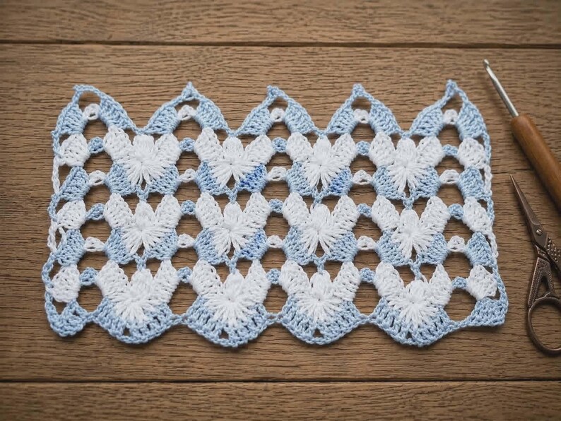 May include: A rectangular piece of crochet lace featuring a repeating chevron pattern in alternating white and light blue yarn. The openwork design consists of star-like clusters and scalloped edges, accompanied by a wooden crochet hook and scissors.
