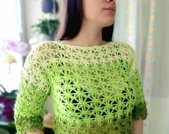 Pode incluir: Um top de crochê verde com um padrão floral. O top de manga curta apresenta um gradiente de verde claro a escuro. A gola e as mangas têm uma borda ondulada. O top é usado sobre uma peça de roupa preta.