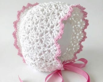 Pode incluir: Um gorro de bebê de crochê branco com uma borda rosa ondulada e uma fita de cetim rosa combinando. O gorro é feito à mão com um design de tecido aberto.