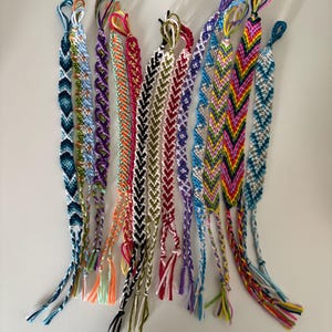 May include: A collection of twelve handmade friendship bracelets crafted from colourful embroidery floss. Each bracelet features intricate woven patterns, including chevron and diagonal stripes in various colour combinations like blue, pink, green, and black.