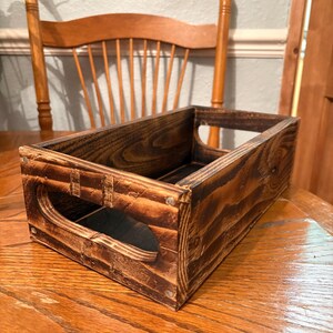 May include: A dark stained wooden crate with cut-out handles. The rectangular box shows visible nail heads and prominent wood grain. The crate is placed on a wooden surface, showcasing its rustic design.
