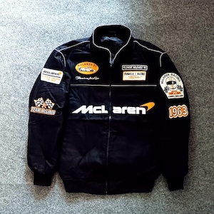 Può includere: Una giacca nera con zip e branding McLaren Formula 1. La parte anteriore mostra il logo McLaren, mentre le toppe includono un'auto da corsa, bandiere a scacchi, il testo 1963 e coordinate. Il design presenta profili bianchi e polsini a coste.
