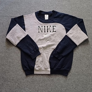 May include: A long-sleeve crewneck sweatshirt featuring a color-blocked design in navy blue and light gray. The center panel is light gray, while the sleeves and sides are navy blue. The word NIKE is embroidered in white with a black outline.