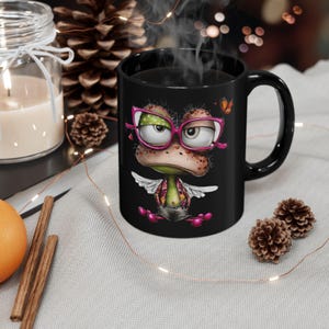 Puede incluir: Taza de cerámica negra con un diseño de rana de dibujos animados. La rana lleva gafas rosas y un chaleco estampado. La taza está llena de un líquido oscuro, posiblemente té o café.