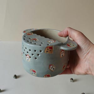 May include: A light blue ceramic berry bowl with two side handles and a series of drainage holes on one side. The exterior surface is decorated with small, hand-painted illustrations of houses featuring red roofs and white walls scattered throughout.