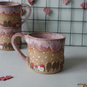 May include: A ceramic mug with a light brown base, adorned with white hearts and a village scene of houses and trees. The rim and handle are a soft pink, with a textured, speckled appearance. The mug is approximately 4 inches tall.