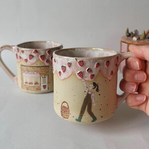 May include: Ceramic mugs with a whimsical strawberry theme. The mugs have a beige base, pink handles, and a strawberry-decorated rim. One mug features a shop illustration, while another depicts a person picking strawberries. A basket of strawberries is also shown.
