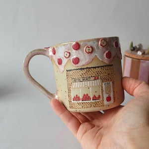 May include: A handmade ceramic mug with a whimsical apple theme. The mug features a tan base with a decorative apple house design, red apple accents, and the text "APPLE HOUSE" and "HOLA". The handle and rim have a pinkish hue.