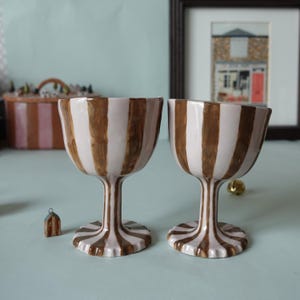 May include: Two ceramic goblets with a white and brown striped pattern. The goblets have a stem and a wide base. A small decorative house and a framed picture are in the background. The goblets are likely for serving beverages.