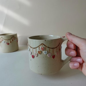 May include: A speckled ceramic mug with a heart garland design. The mug has a handle and is decorated with red, pink, and patterned hearts. Another similar mug is in the background.