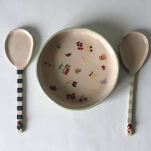 Puede incluir: Un cuenco de cerámica y dos cucharas de servir. El cuenco es beige con un borde verde y está decorado con ilustraciones coloridas con temática veraniega. Las cucharas tienen cabezas beige y mangos a rayas verdes y blancas, con un detalle de mariquita.
