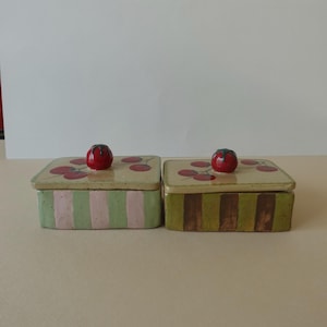 Könnte beinhalten: Zwei rechteckige Keramikboxen mit Deckel. Die Deckel sind beige mit roten Kirschmotiven. Jede Box hat einen roten Kirschknauf. Eine Box hat grüne und rosa Streifen, die andere grüne und braune Streifen.