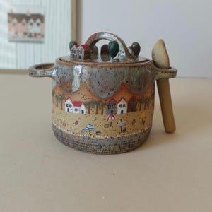 May include: A speckled ceramic jar with a lid featuring miniature house sculptures and a matching spoon. The jar body displays a painted beach scene with small houses, palm trees, and colourful umbrellas, finished with a scalloped top edge.