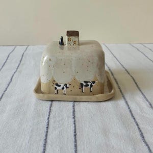 May include: A ceramic butter dish with a lid, featuring a whimsical design. The dish is beige with a speckled pattern and a painted scene of two cows. A small house and chimney are on top of the lid. The dish sits on a matching tray.