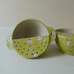 May include: Two lime green ceramic berry bowls with speckled glaze and small handles. The exterior features a repeating pattern of tiny hand-painted houses. Each bowl has a series of drainage holes on the front side for rinsing fruit.