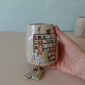 May include: A speckled beige ceramic mug featuring a hand-painted illustration of a person with red hair reading a book in a chair next to a tall bookshelf filled with colourful books, plants, and a clock. A matching ceramic charm is attached below.
