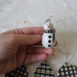 Pode incluir: Um pequeno enfeite de boneco de neve branco com um cachecol azul e dourado, olhos de botão pretos e um nariz de cenoura laranja. O boneco de neve é segurado por um fio dourado, sugerindo que é para pendurar. Decoração de Natal.