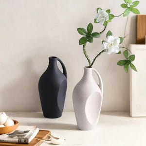 May include: Two ceramic vases, one black and one white, with a single stem of white flowers in the white vase. The vases have a unique, modern design with a handle on the side.