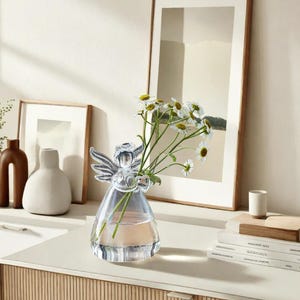 Puede incluir: Un jarrón de cristal transparente con forma de ángel con un ramo de flores blancas. El jarrón está sobre una encimera blanca con un armario de madera debajo.