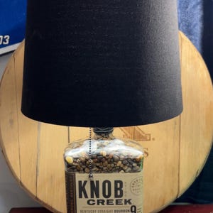 May include: A unique lamp made from a repurposed Knob Creek bourbon whiskey bottle. The lamp features a black lampshade and is filled with small stones. The base is a wooden circle, and the lamp is on a red surface.