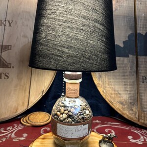 May include: A unique lamp made from a repurposed bottle filled with small stones. The lamp features a black lampshade and sits on a wooden base. The bottle has a label with handwritten text. A small metal figurine is on the base.