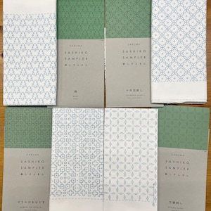 May include: Eight different sashiko sampler fabric panels with a green background and blue stitching. The panels feature different geometric patterns, including a cross stitch, a diamond pattern, and a tree pattern. The panels are labeled with the Japanese words "Sashiko Sampler" and "Daruma".