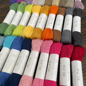 以下が含まれることがあります： 様々な色合いの綿刺繍糸が24個、糸巻きに巻かれた状態で並んでいます。糸巻きには「刺し子糸」と記載されています。糸巻きは木製の台の上に並べられています。糸の色は、緑、青、ピンク、赤、オレンジ、黄、茶色などがあります。