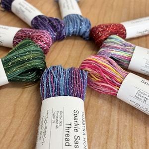 May include: A circular arrangement of eight skeins of Hobbyra Hobbyre Sparkle Sashiko thread in various colours including purple, blue, red, green, and multicoloured blends. Each skein features a white paper label with text detailing fibre content.