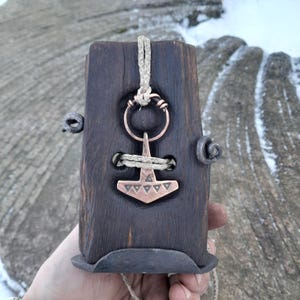 May include: A dark brown wooden block with a copper-coloured pendant in the shape of a hammer. The pendant is attached with a beige rope. The block has metal accents and is held in a hand.